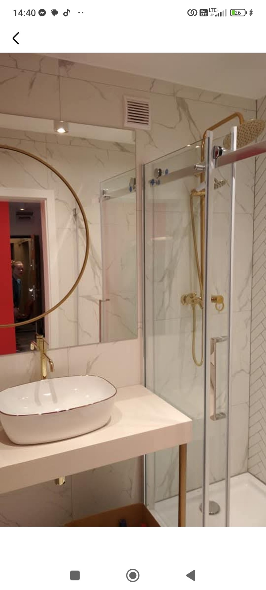 Nowoczesna łazienka z kabiną prysznicową typu walk-in, umywalką nablatową i okrągłym lustrem z złotymi akcentami, marmurowe płytki na ścianach.