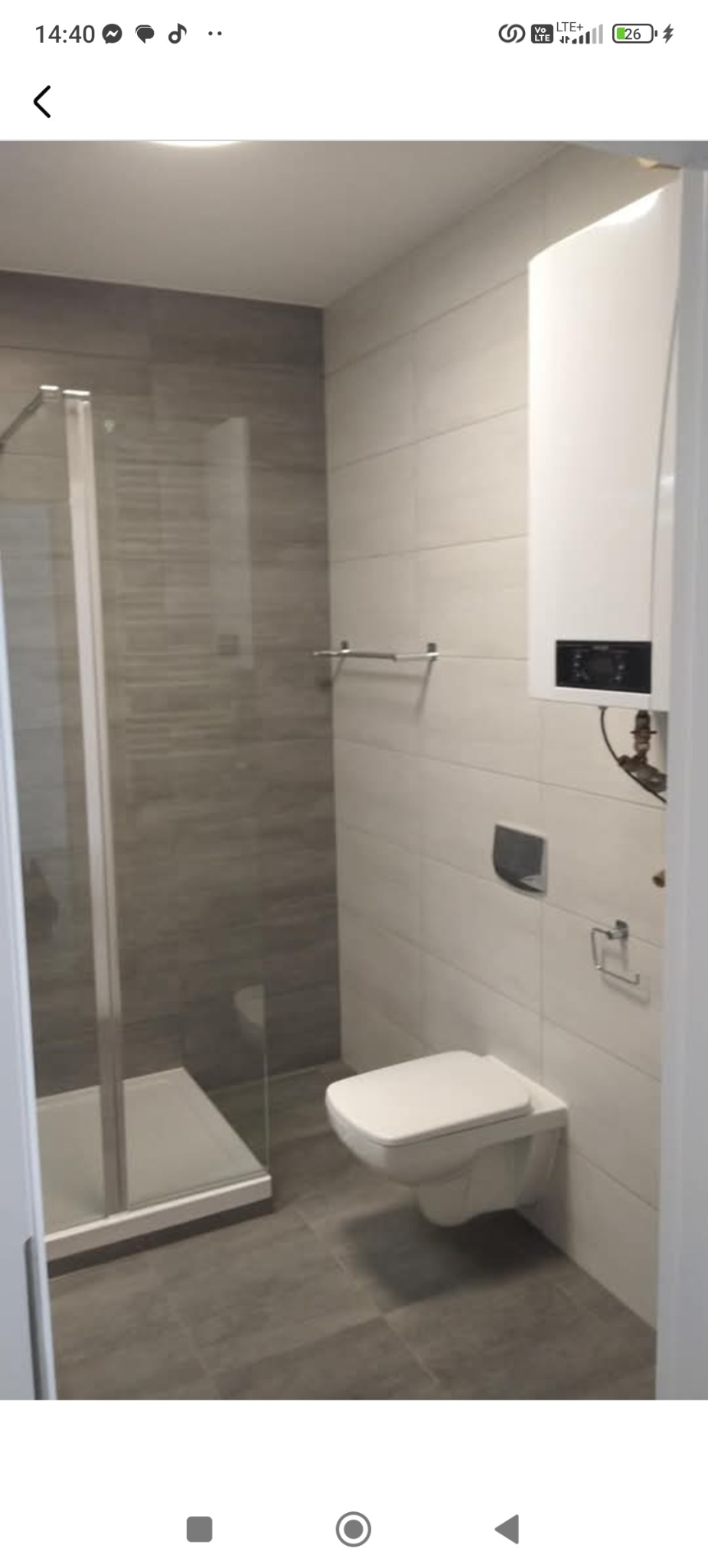 Nowoczesna łazienka po remoncie: kabina prysznicowa ze szklanymi drzwiami, podwieszana toaleta, grzejnik i szare płytki podłogowe oraz ścienne.