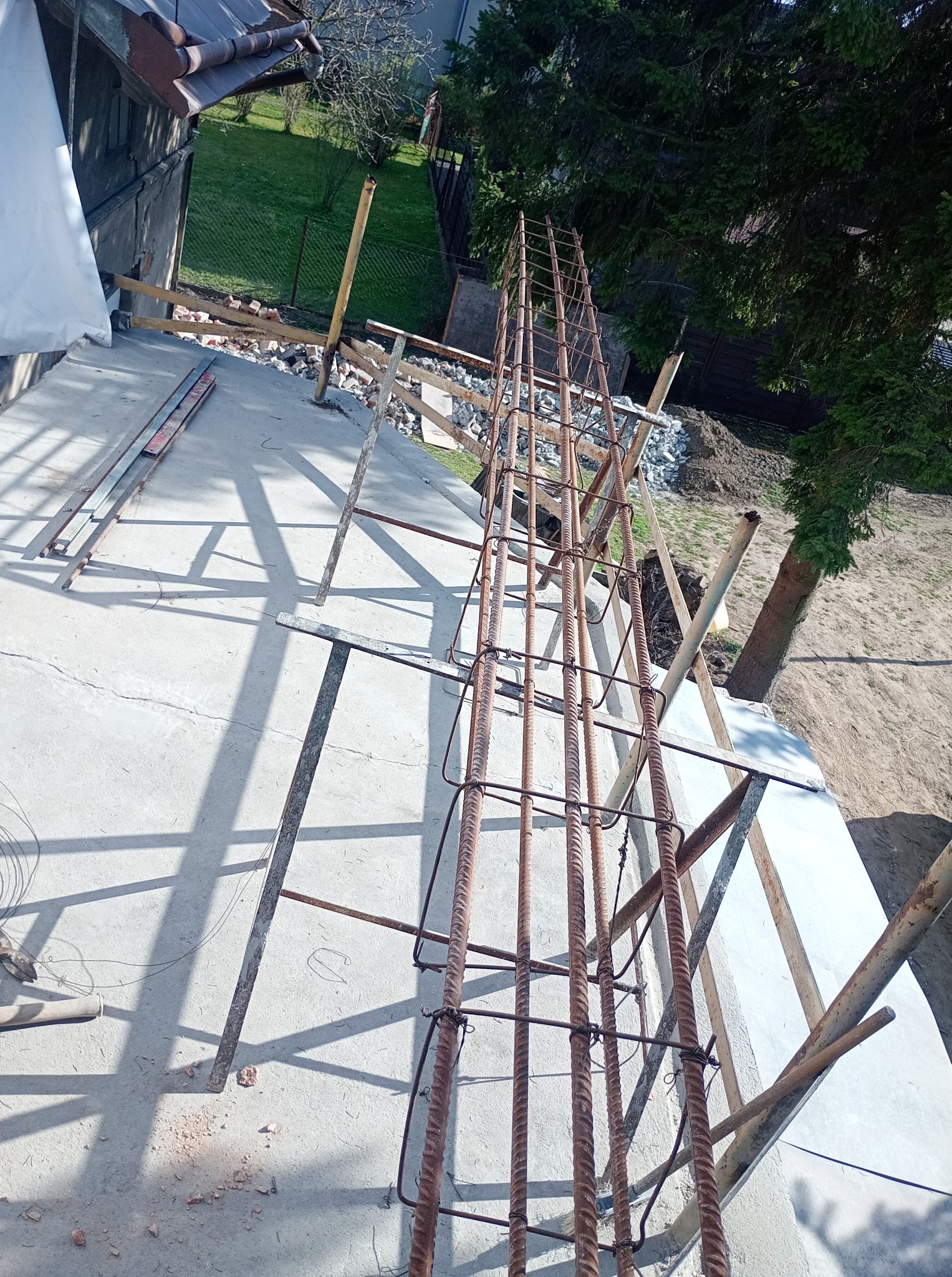 Zbrojenie pionowe na budowie domu jednorodzinnego. Widoczne pręty zbrojeniowe i elementy szalunku. Kąt ujęcia z góry, w tle zieleń i fragment dachu.
