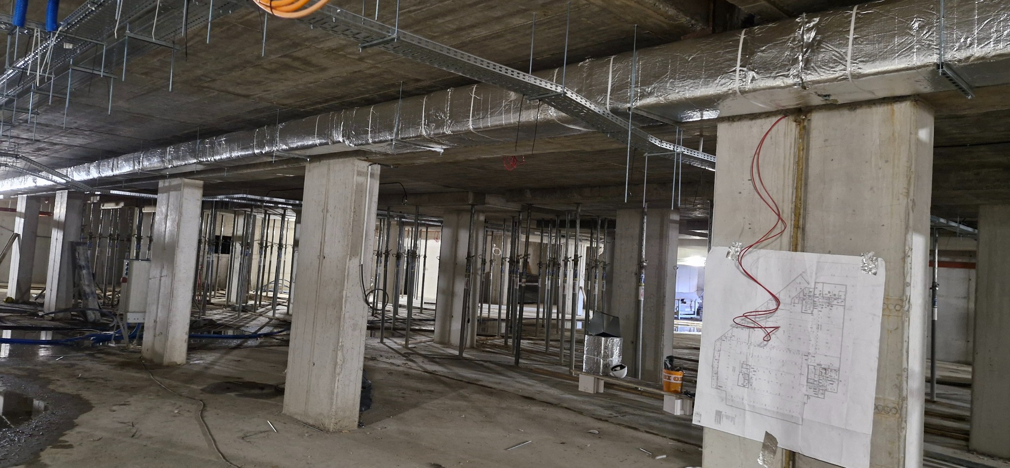 Surowy stan: instalacja wentylacji w budynku. Widoczne kanały z izolacją, koryta kablowe, betonowe słupy i plan techniczny na słupie.