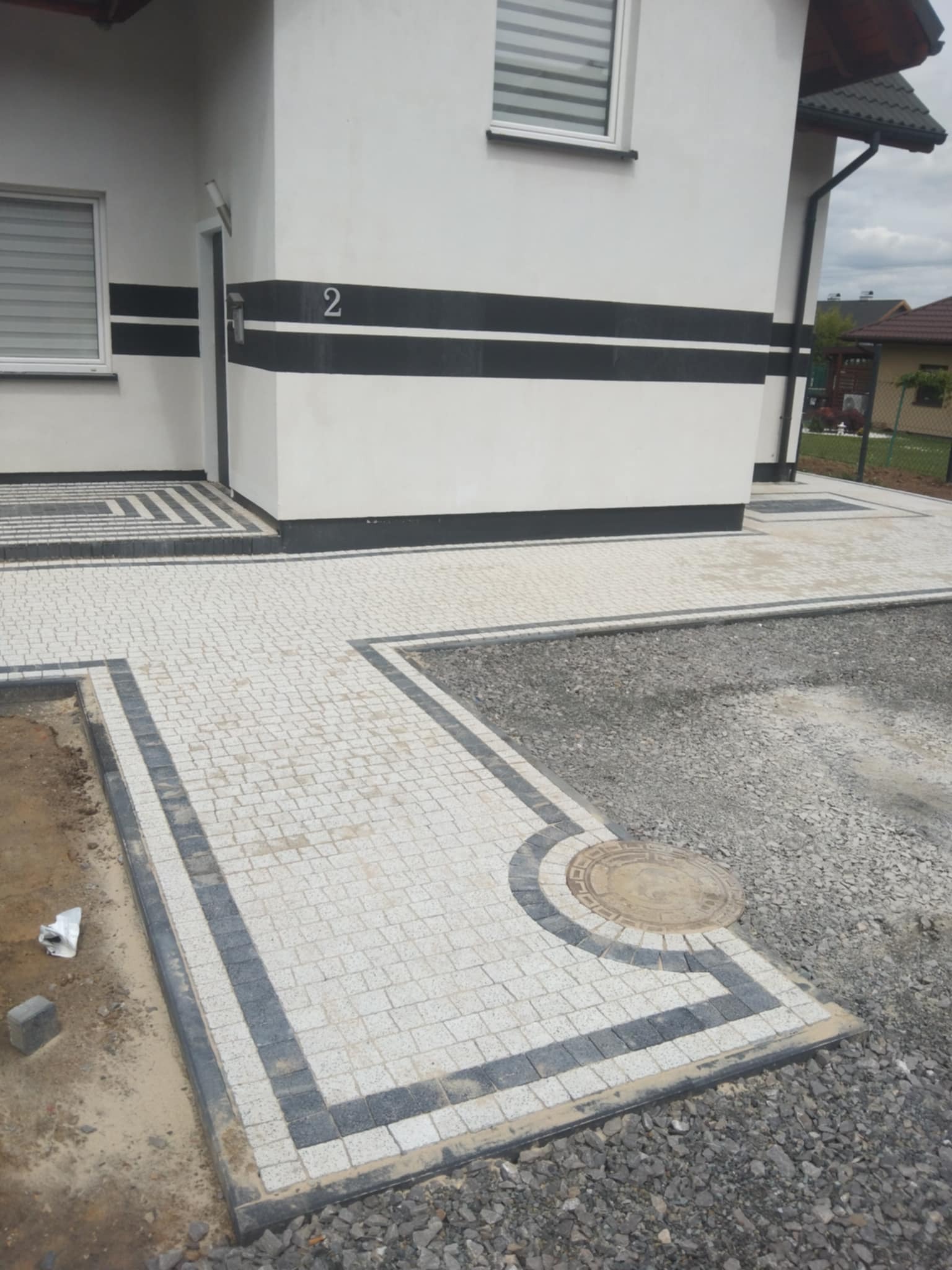 Biało-szara kostka brukowa ułożona wzdłuż ściany budynku z numerem 2, obrzeża z ciemnej kostki. Częściowo wysypany żwir. Widok na fragment posesji.