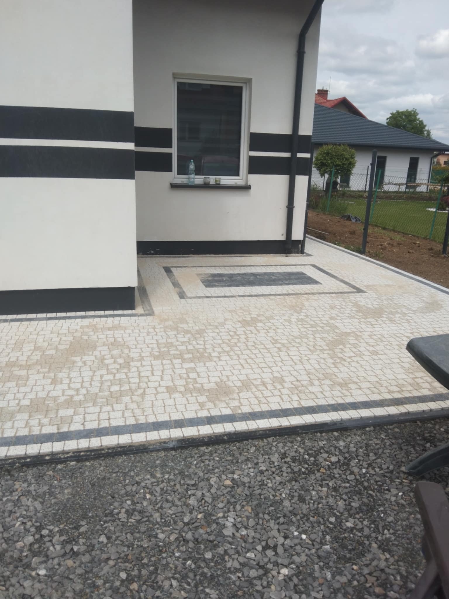 Ułożona jasna kostka brukowa z ciemnymi obramowaniami, tworząca wzór przed budynkiem z oknem. Widoczne szare kamienie na obrzeżach.
