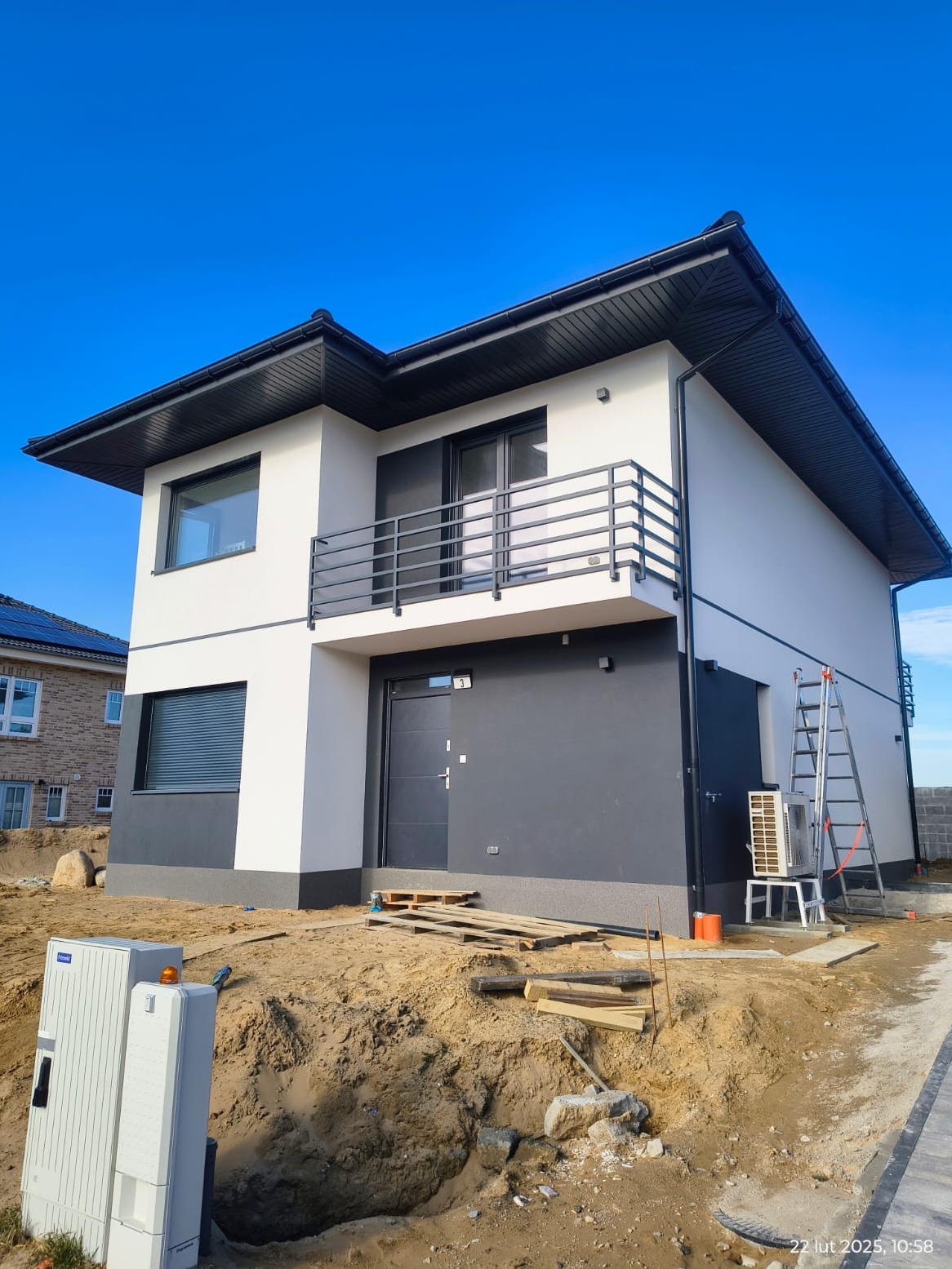 Nowoczesny dom jednorodzinny z balkonem i szarą stolarką okienną, widziany z dołu pod błękitnym niebem. Teren wokół domu w trakcie prac wykończeniowych.