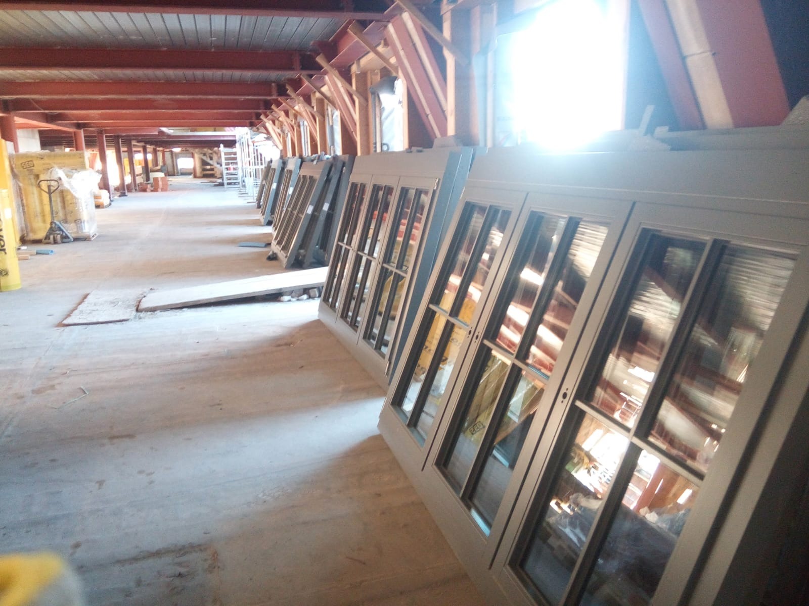 Rząd nowych okien z szybami, ustawionych w surowym wnętrzu budynku w trakcie budowy, z widoczną konstrukcją dachu i materiałami budowlanymi w tle.