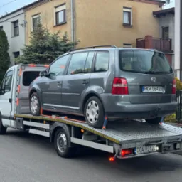 Laweta z szarym VW Touran, zabezpieczonym pasami, na tle miejskiej zabudowy. Widoczne tablice rejestracyjne pojazdów i detale konstrukcyjne lawety.