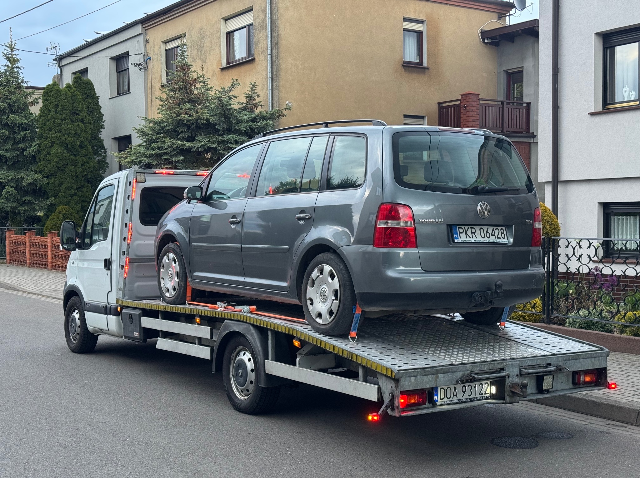 Laweta z szarym VW Touran, zabezpieczonym pasami, na tle miejskiej zabudowy. Widoczne tablice rejestracyjne pojazdów i detale konstrukcyjne lawety.