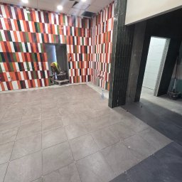 Krzysztof Wsolak - Wnętrze w trakcie prac glazurniczych: ułożone płytki podłogowe i wzorzyste płytki ścienne w odcieniach czerwieni, pomarańczy, zieleni i bieli. Widoczny pracownik w tle.