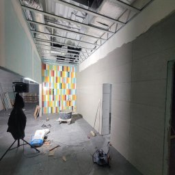 Krzysztof Wsolak - Wnętrze w trakcie prac wykończeniowych: ułożona szara podłoga, częściowo położone płytki na ścianach, kolorowa ściana z płytek. Widoczne narzędzia i materiały budowlane.