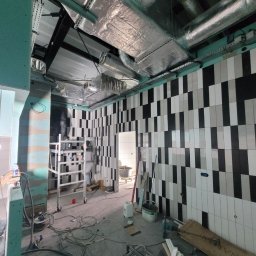 Krzysztof Wsolak - Wnętrze w trakcie prac: ściany z biało-czarną glazurą ułożoną w pasy, widoczne instalacje pod sufitem, narzędzia i materiały budowlane. Scena z budowy/remontu.