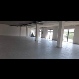 Krzysztof Wsolak - Wnętrze dużego, jasnego pomieszczenia biurowego z szarą podłogą i białymi kolumnami. Widoczne okna i pracownik w oddali. Przestrzeń przygotowana do wykończenia.