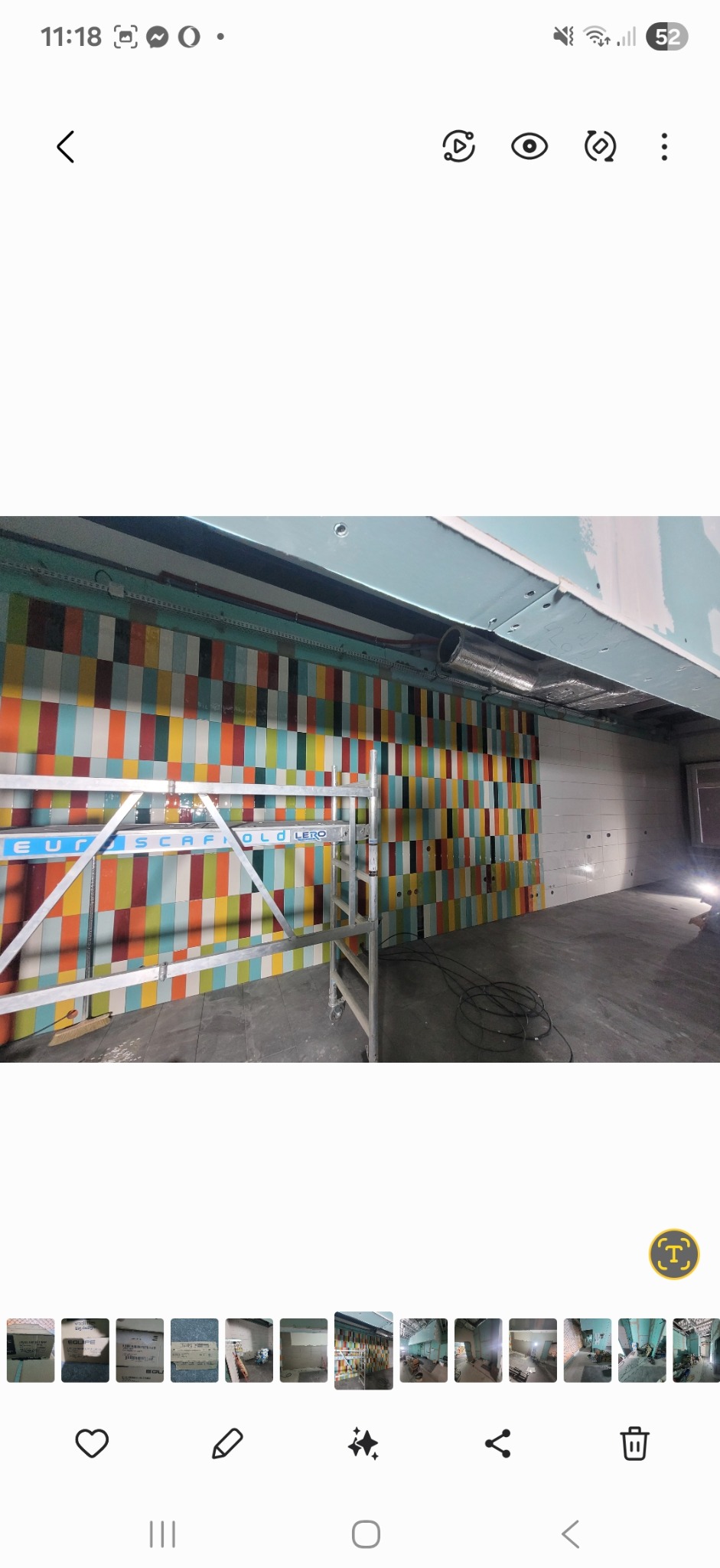 Ściana z kolorowymi płytkami w trakcie układania, widoczne rusztowanie. Część ściany wyłożona białymi płytkami. Sufit w trakcie wykończenia płytami gipsowymi.