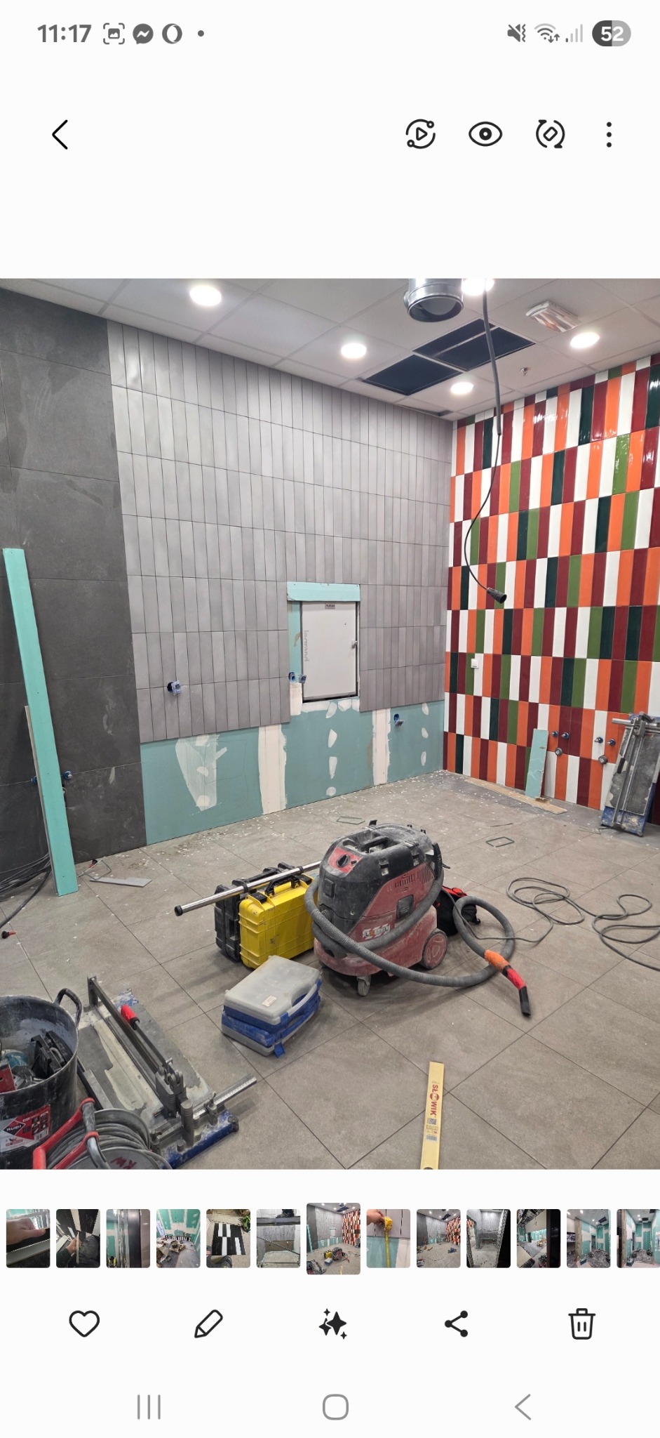 Wnętrze w trakcie kładzenia płytek na ścianach, widoczne narzędzia glazurnicze i odkurzacz przemysłowy. Częściowo wykończone ściany z różnymi wzorami płytek.