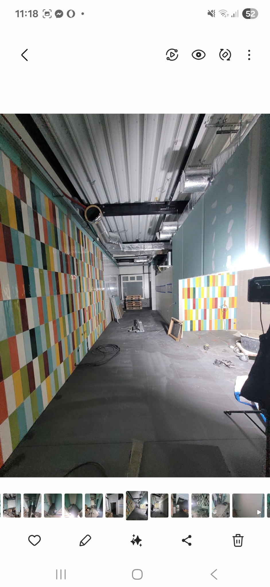 Wnętrze w trakcie remontu: ściana z kolorową glazurą w pionowe pasy, szara podłoga, widoczne instalacje pod sufitem. Częściowo wykończone pomieszczenie.