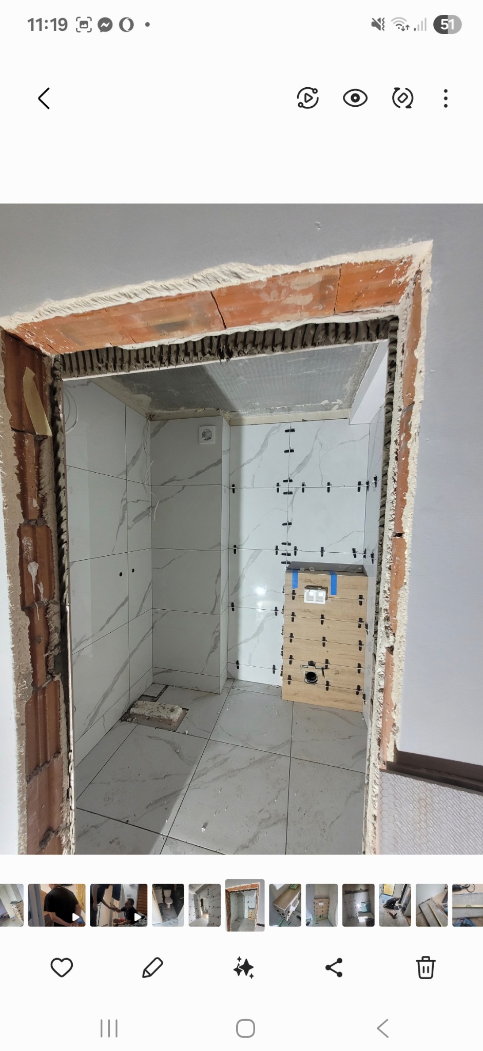 Wnętrze łazienki w trakcie remontu. Widoczne białe płytki imitujące marmur na ścianach i podłodze, krzyżyki dystansowe i obudowa stelaża podtynkowego.