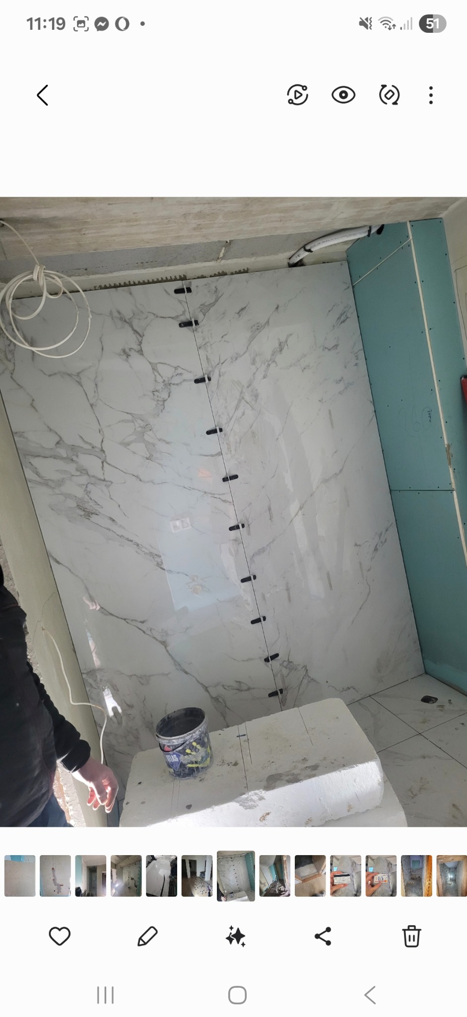 Układanie płytek imitujących marmur na ścianie z użyciem klipsów poziomujących. Widoczna ściana z płyt kartonowo-gipsowych i wiadro po zaprawie.