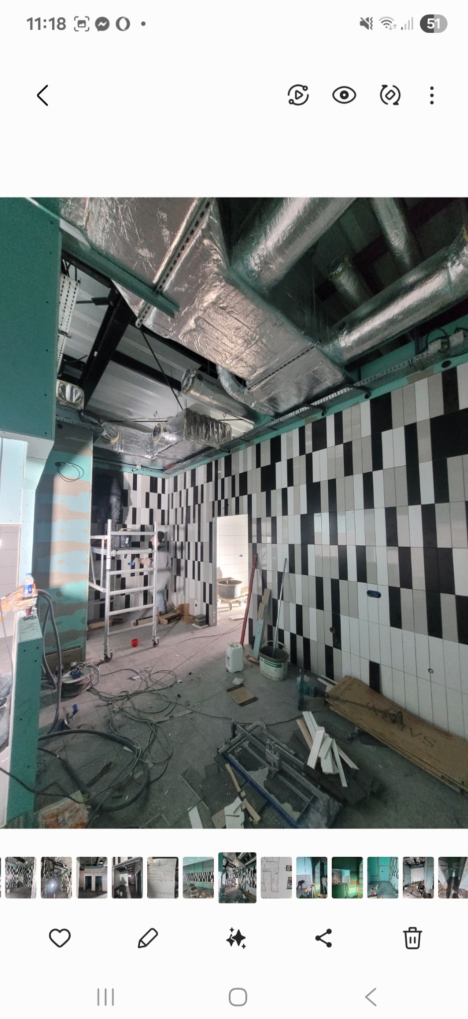 Wnętrze w trakcie prac: ściany z biało-czarną glazurą ułożoną w pasy, widoczne instalacje pod sufitem, narzędzia i materiały budowlane. Scena z budowy/remontu.