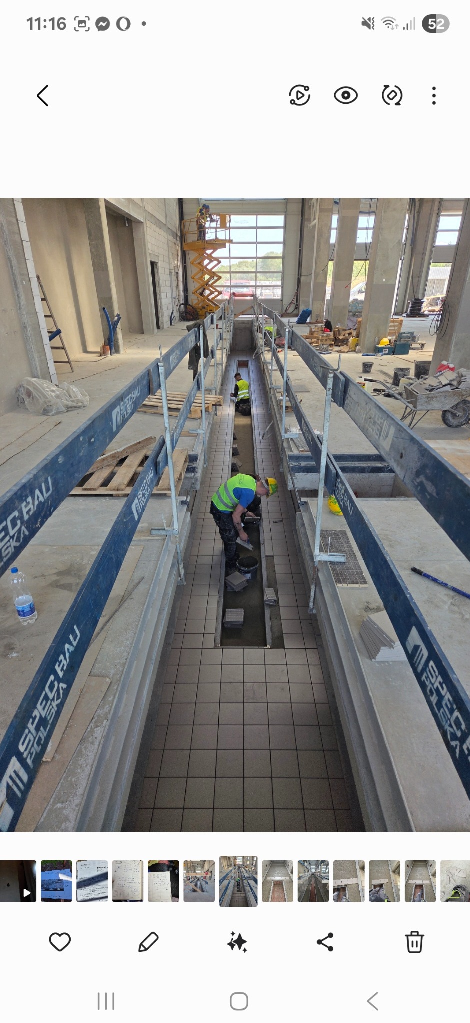 Układanie szarych płytek podłogowych w długim kanale. Pracownicy w kamizelkach odblaskowych i kaskach. Spec Bau Polska na barierkach. Hala przemysłowa w tle.