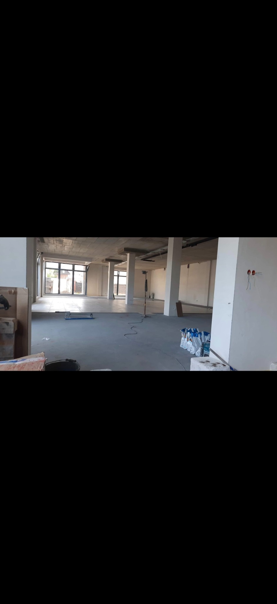 Wnętrze w stanie deweloperskim: surowy beton, kolumny, duże okna. Widoczne materiały budowlane i instalacje elektryczne. Przestrzeń przygotowana do wykończenia.