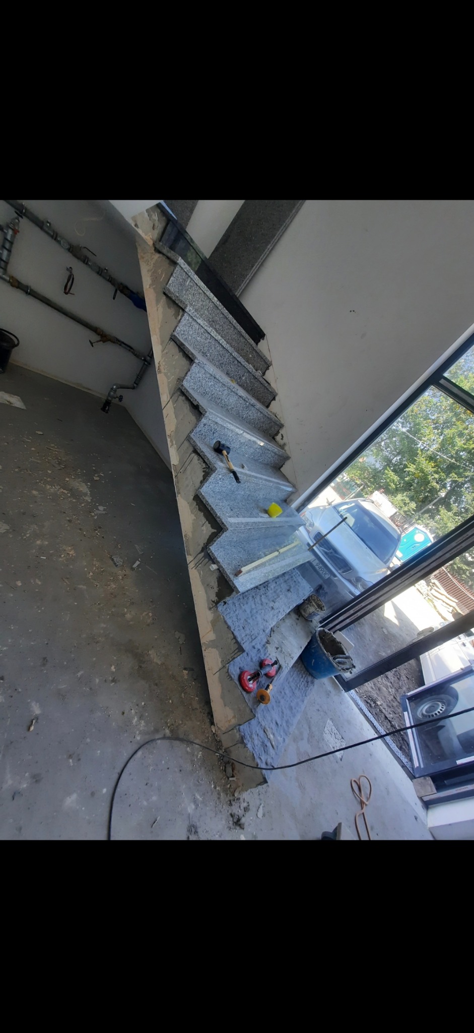 Układanie szarych płytek na schodach wewnętrznych. Widoczne narzędzia glazurnicze, młotek i przyssawki. Prace wykończeniowe w trakcie realizacji.