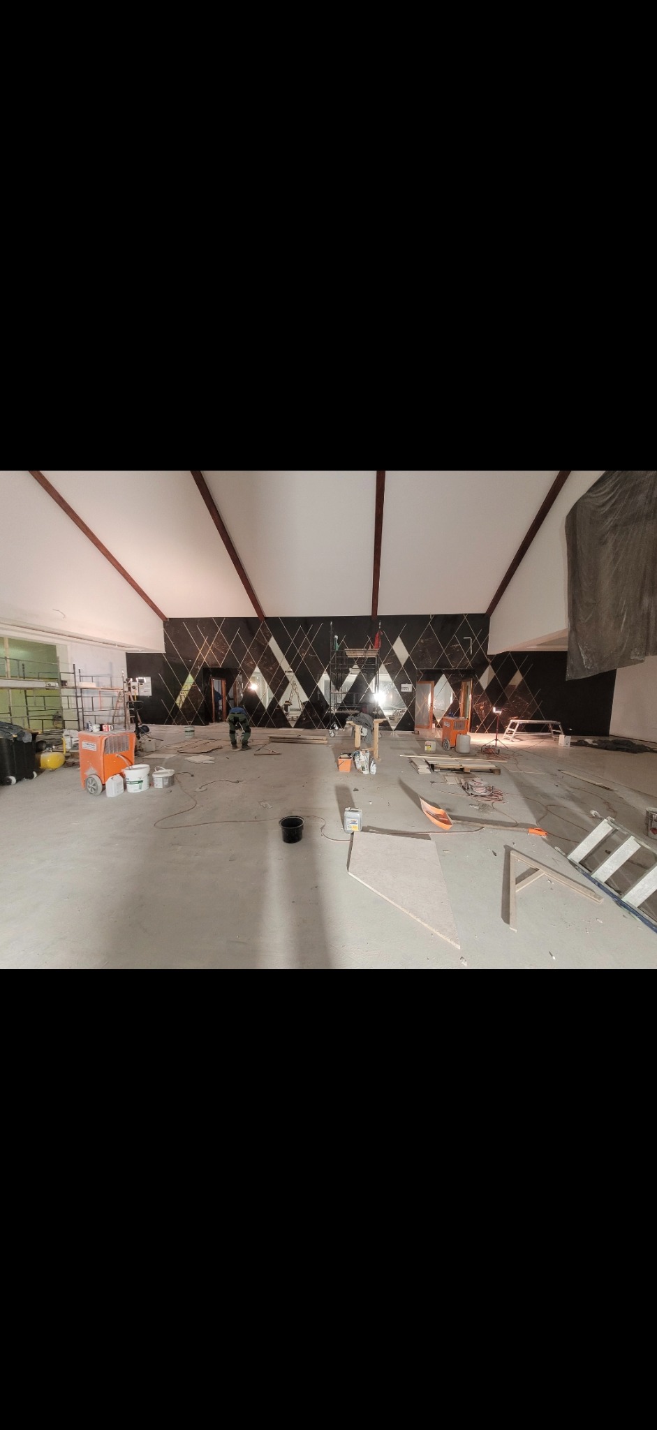 Duże pomieszczenie w trakcie wykańczania, ściana z dekoracyjnymi panelami w romby, widoczne narzędzia i materiały budowlane, dwóch pracowników w oddali.