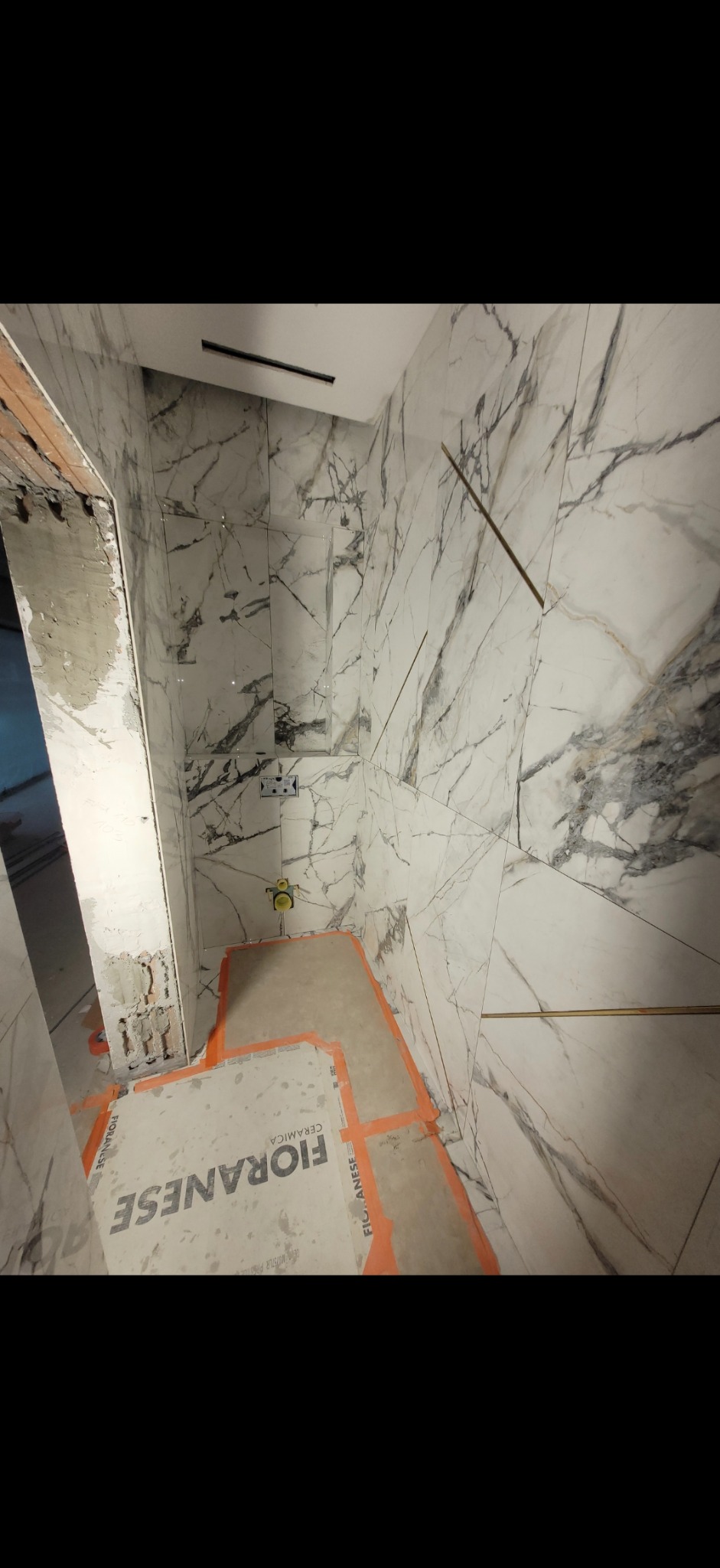 Łazienka w trakcie wykończenia: marmurowe płytki na ścianach, widoczne elementy instalacji i zabezpieczone podłoże. Prace glazurnicze w toku.