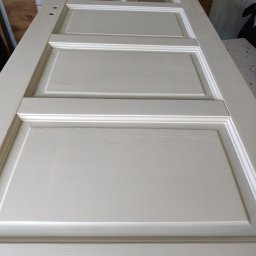 S&M BEST - Białe, odnowione drzwi z szybkami, leżące na płasko. Widoczne otwory na klamkę i zamek. Świeżo pomalowana powierzchnia z delikatną strukturą drewna.