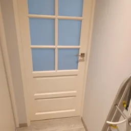 Białe drzwi wewnętrzne z szybami w kolorze błękitu, widoczne na tle jasnej ściany i podłogi. Fragment drabiny malarskiej w prawym dolnym rogu.