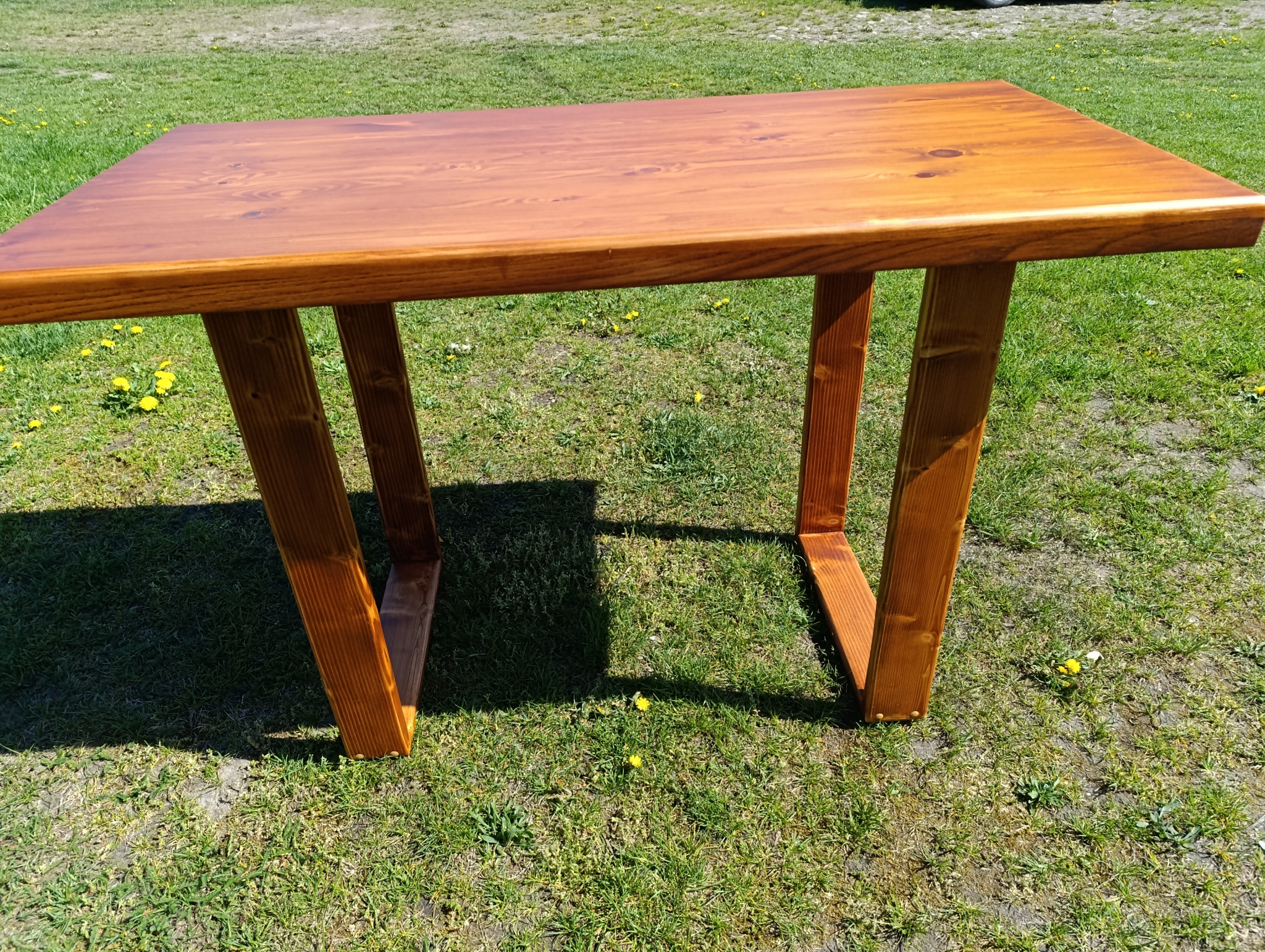 Stół drewniany, prostokątny, o gładkim blacie i charakterystycznych nogach w kształcie litery V, stoi na trawie w słoneczny dzień. Widoczne sęki w drewnie.