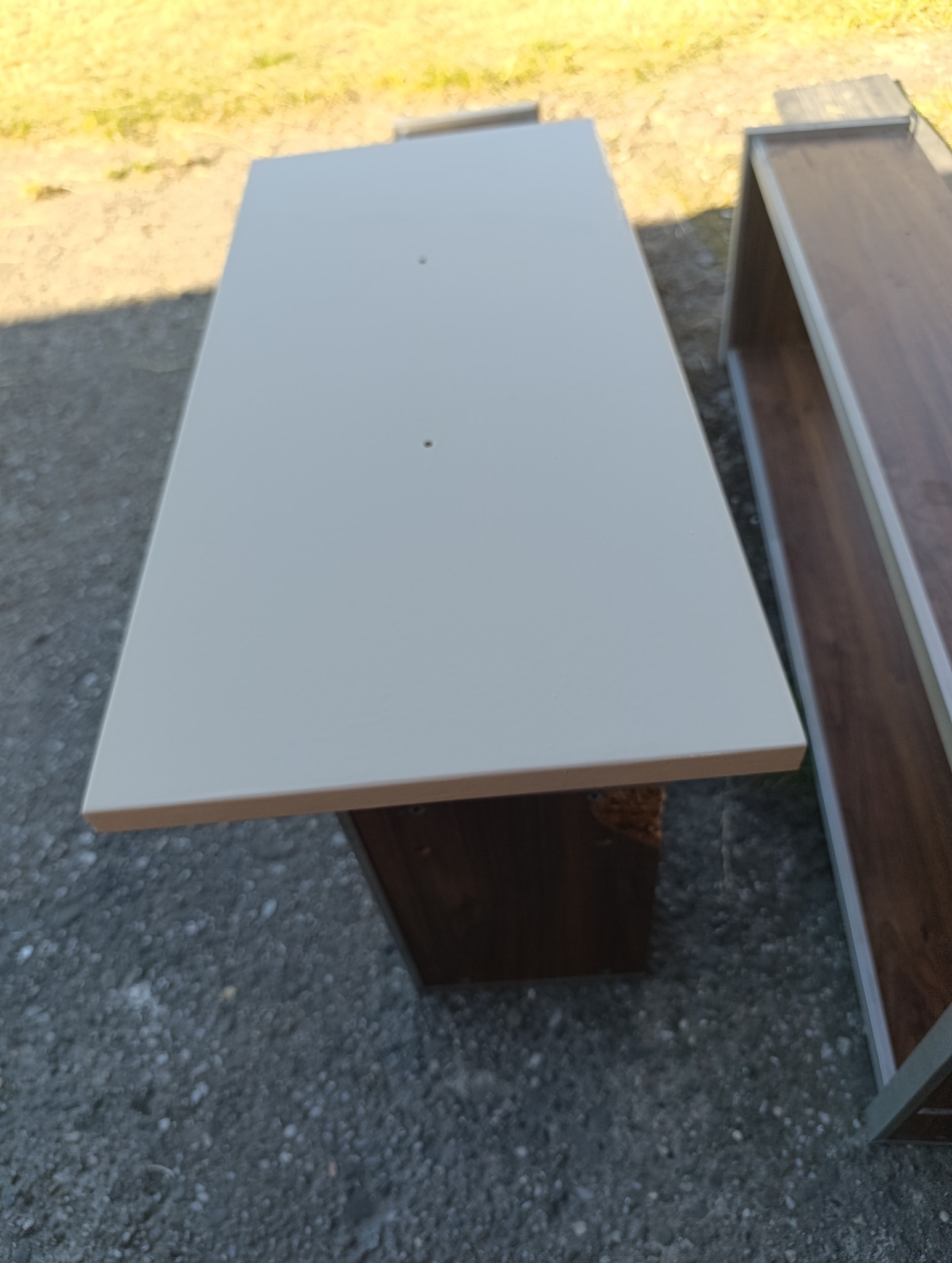 Biały blat stolika z widocznymi otworami na śruby, umieszczony na ciemnej podstawie. Obok fragment brązowego regału. Zdjęcie na szarym, żwirowym podłożu.