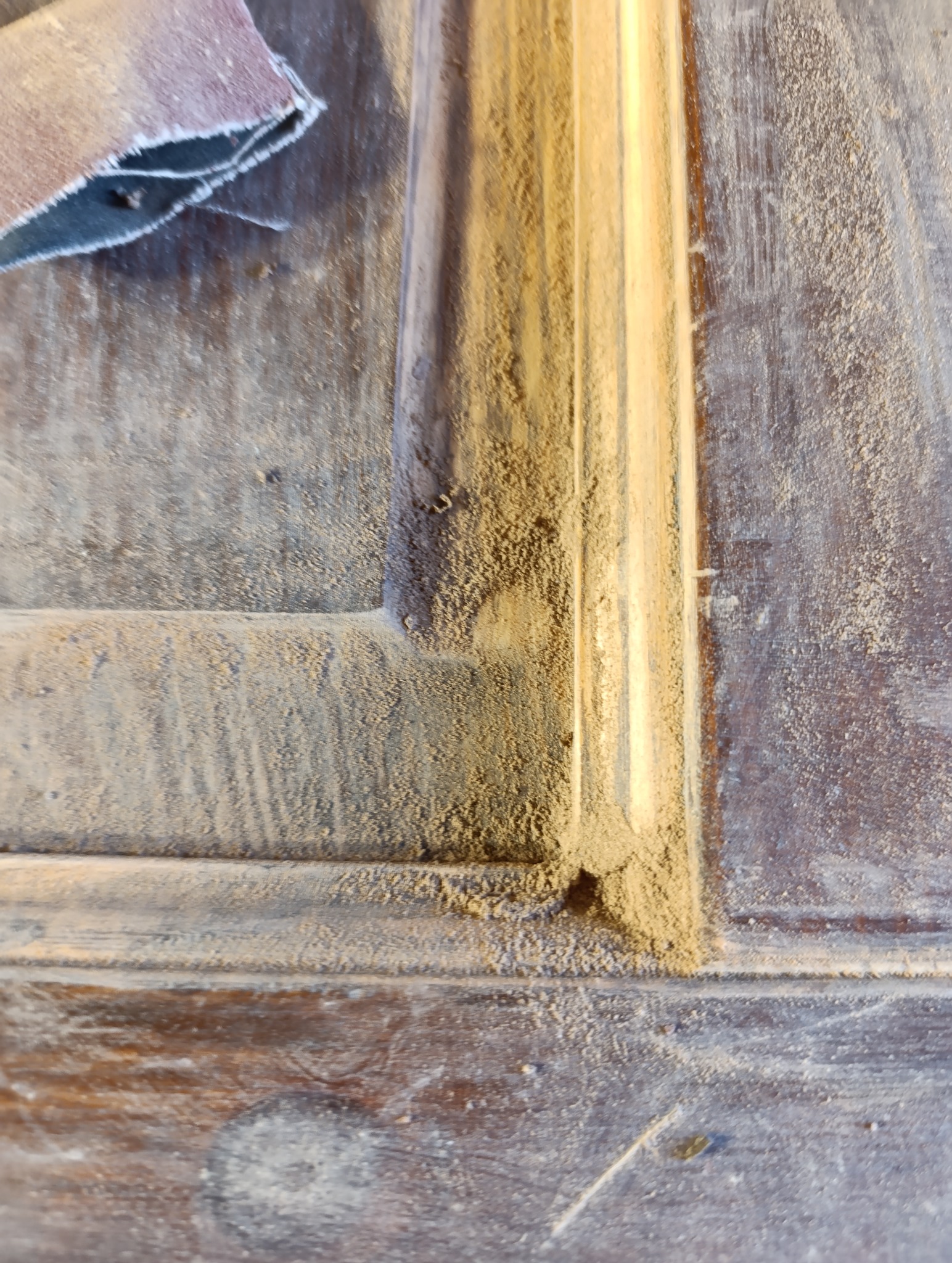 Fragment drewnianej ramy okiennej w trakcie renowacji, pokrytej pyłem po szlifowaniu, z widocznym fragmentem papieru ściernego w lewym górnym rogu.