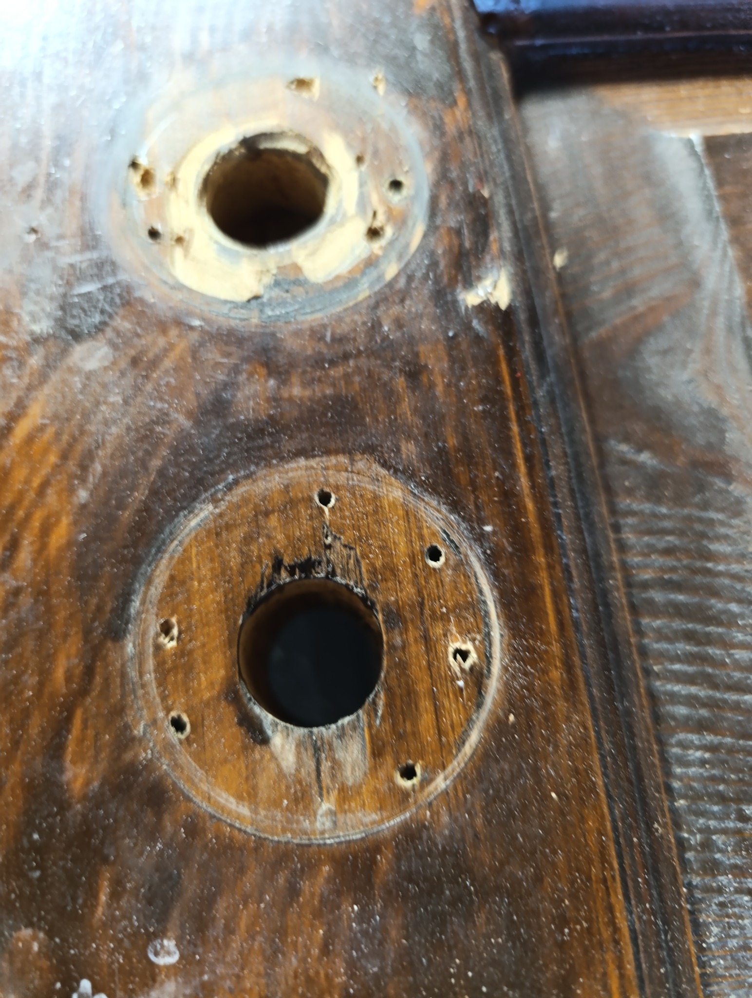 Fragment sfatygowanych, brązowych drzwi drewnianych z dwoma okrągłymi otworami po klamkach i śladami napraw. Widoczne wypełnienia i nawiercenia.