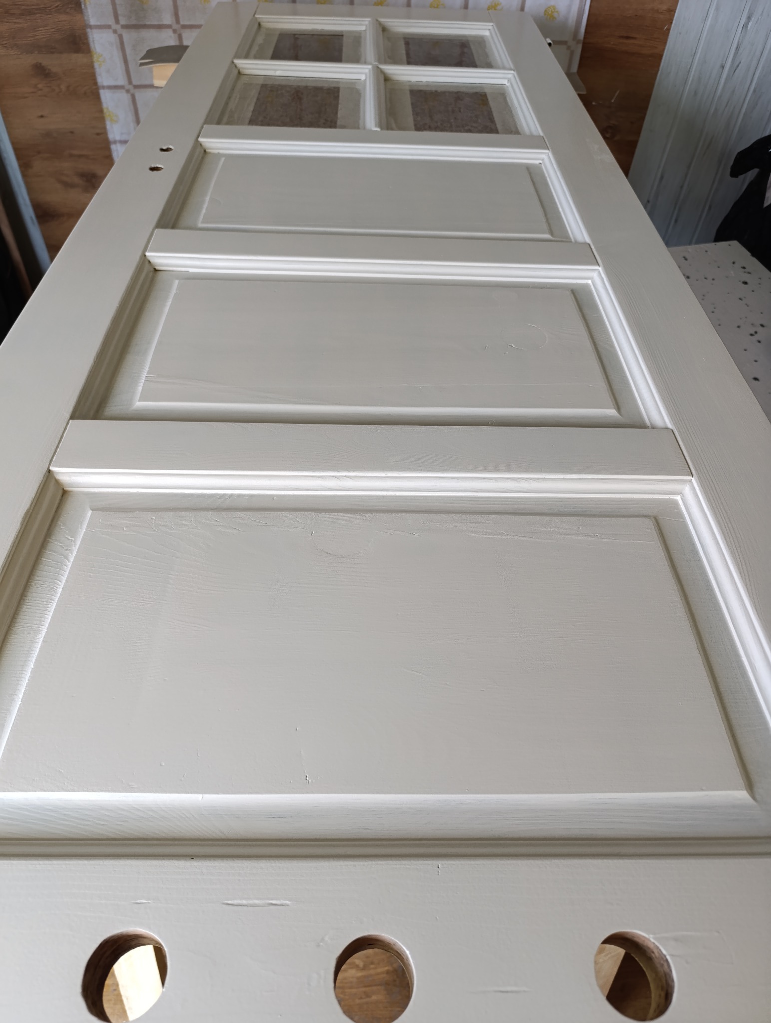 Białe, odnowione drzwi z szybkami, leżące na płasko. Widoczne otwory na klamkę i zamek. Świeżo pomalowana powierzchnia z delikatną strukturą drewna.