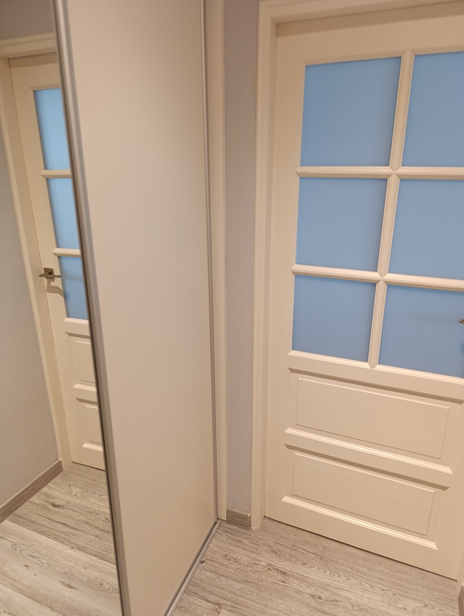 Beżowe drzwi z szybami w kratkę, widoczne obok drzwi przesuwnych szafy. Jasna podłoga. Ujęcie z bliska, prezentujące detale wykończenia.