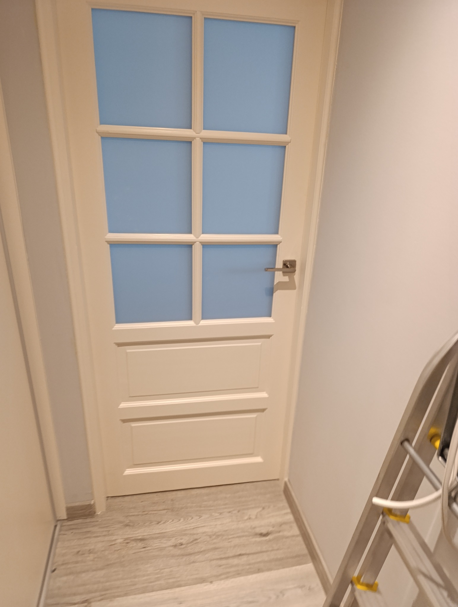 Białe drzwi wewnętrzne z szybami w kolorze błękitu, widoczne na tle jasnej ściany i podłogi. Fragment drabiny malarskiej w prawym dolnym rogu.