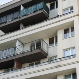 Wielopiętrowy budynek z balkonami zabezpieczonymi siatką ochronną przed ptakami, widok z dołu, neutralne oświetlenie.