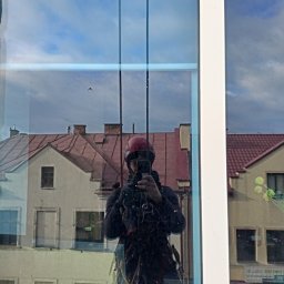 Krzysztof Kalkowski Usługi Wysokościowe - Pracownik w kasku, zabezpieczony linami, robi zdjęcie smartfonem w odbiciu okna budynku. Widoczne budynki w tle i niebo.