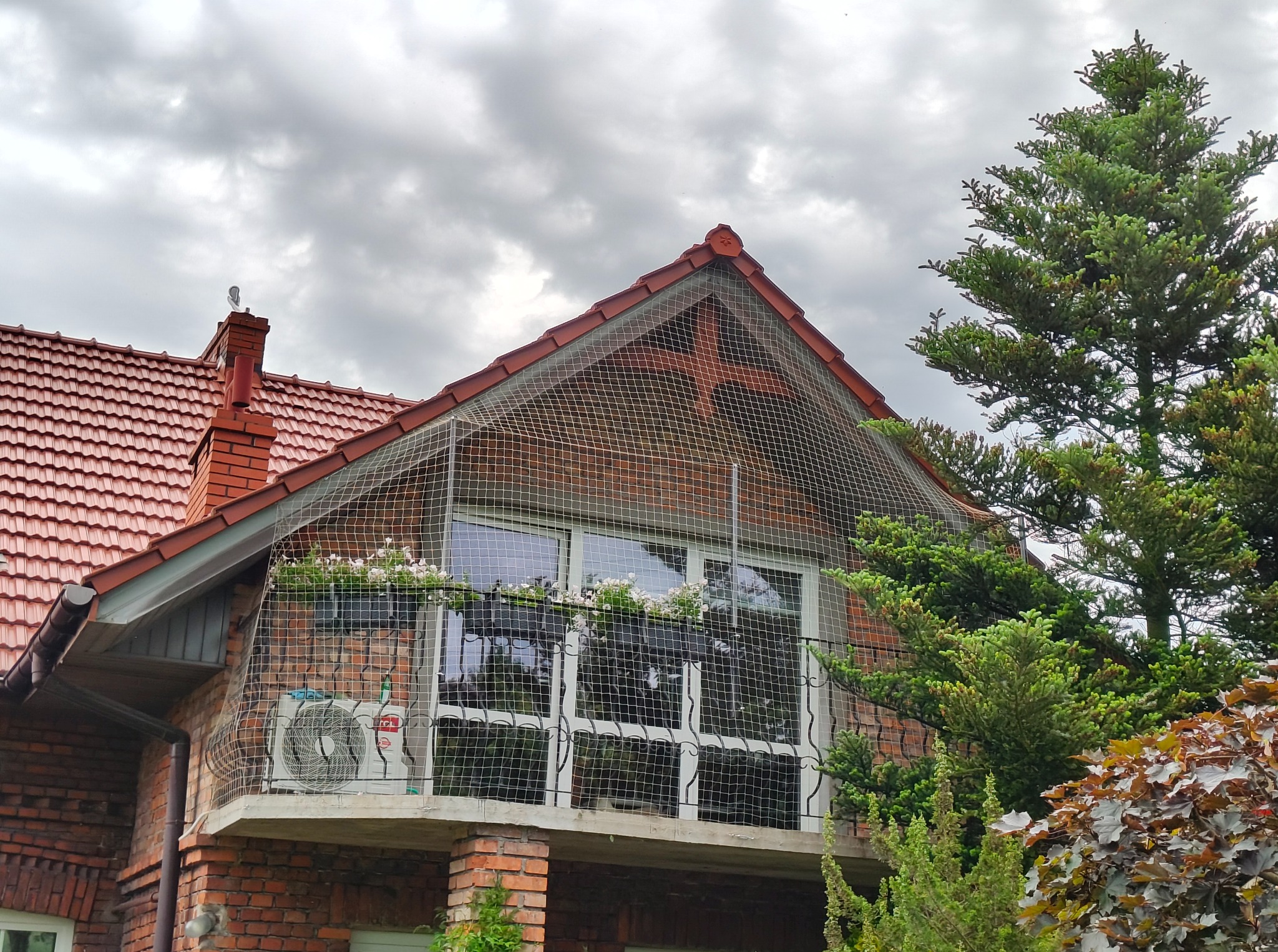 Siatka ochronna na balkonie z cegły chroniąca przed ptakami, z kwiatami w skrzynkach i klimatyzatorem. Czerwona dachówka i zielone drzewa w tle.