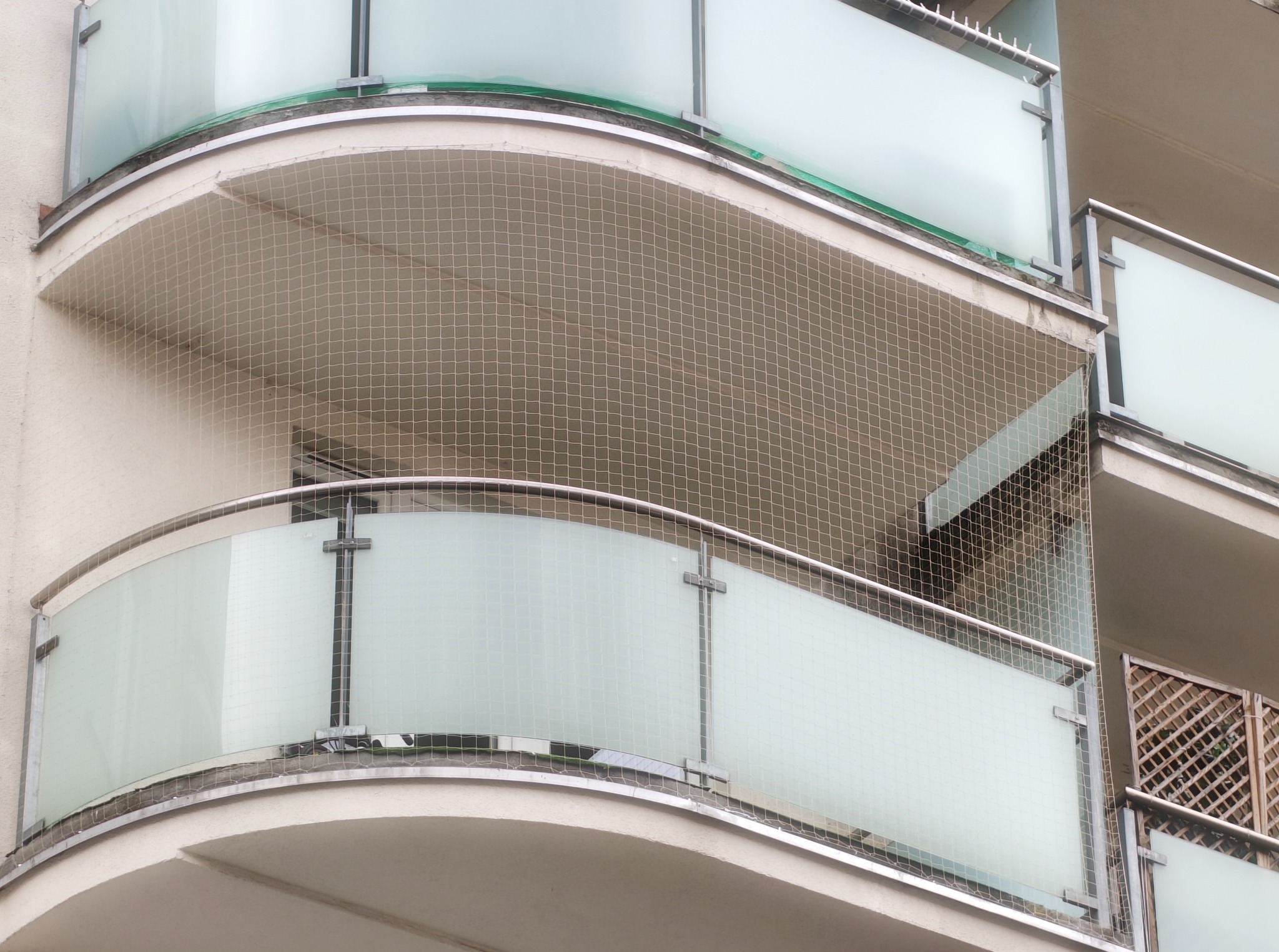 Zabezpieczenie balkonu siatką ochronną przed ptakami, z widocznymi kolcami na górnej krawędzi. Szklane balustrady i metalowe elementy konstrukcyjne.