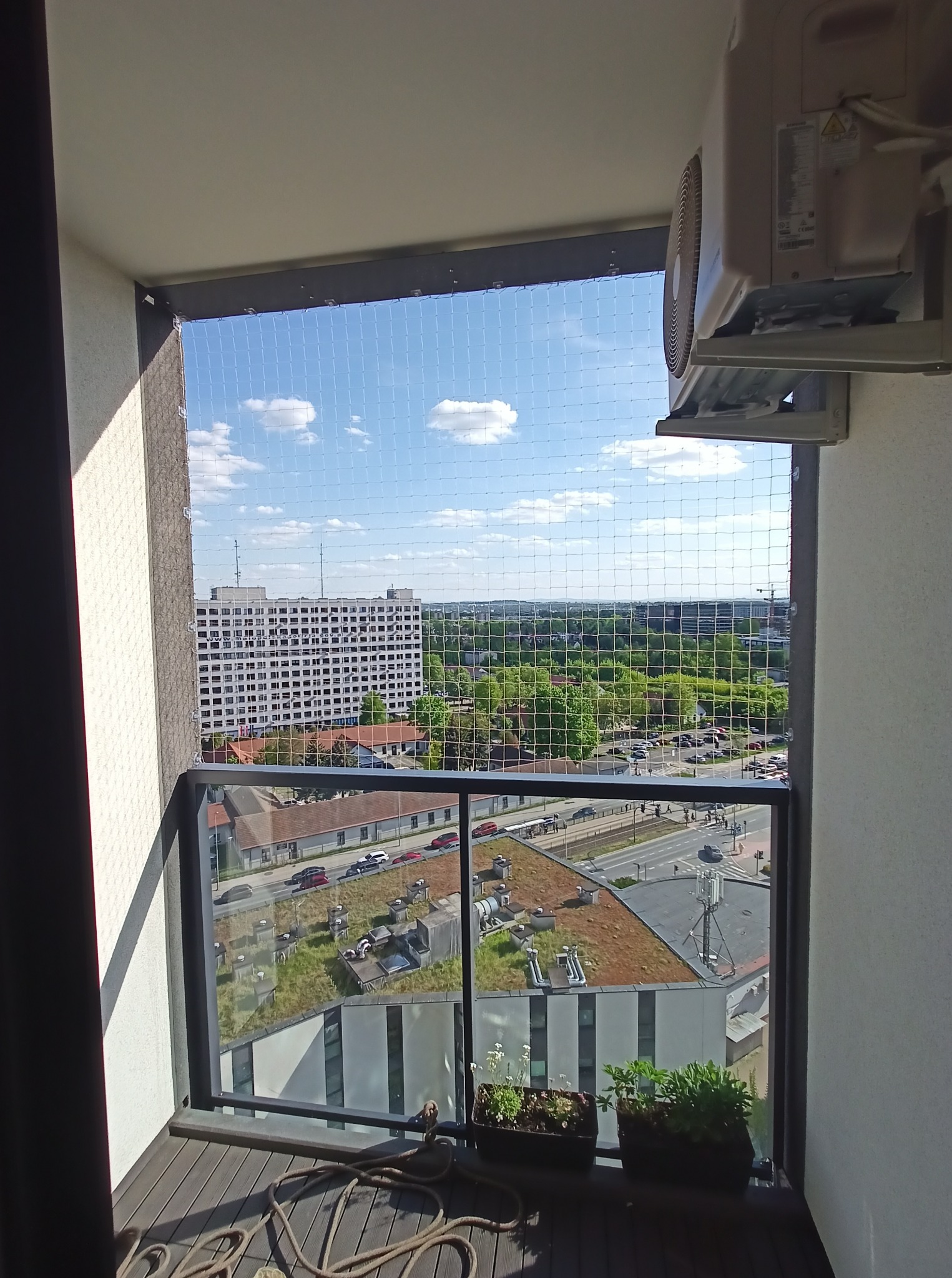 Zabezpieczenie balkonu siatką ochronną z widokiem na miasto. Na balkonie donice z roślinami i zwinięta lina. Widoczny klimatyzator na ścianie.