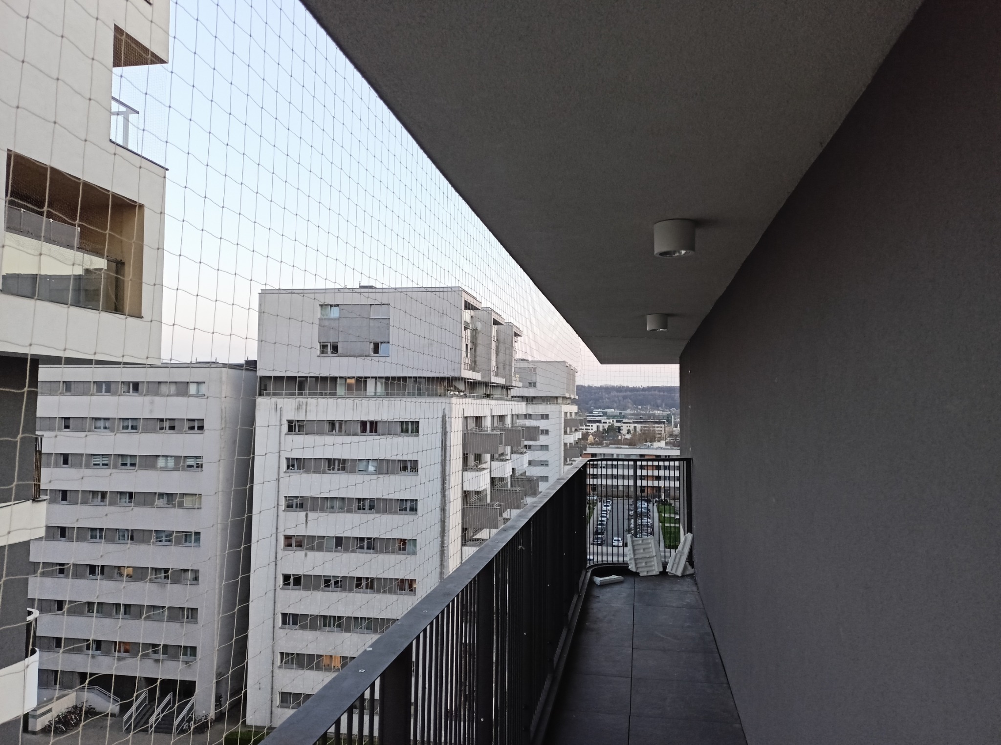 Siatka ochronna na balkonie wysokiego budynku mieszkalnego, zabezpieczająca przed ptakami. Widok na panoramę miasta w tle, z perspektywy balkonu.