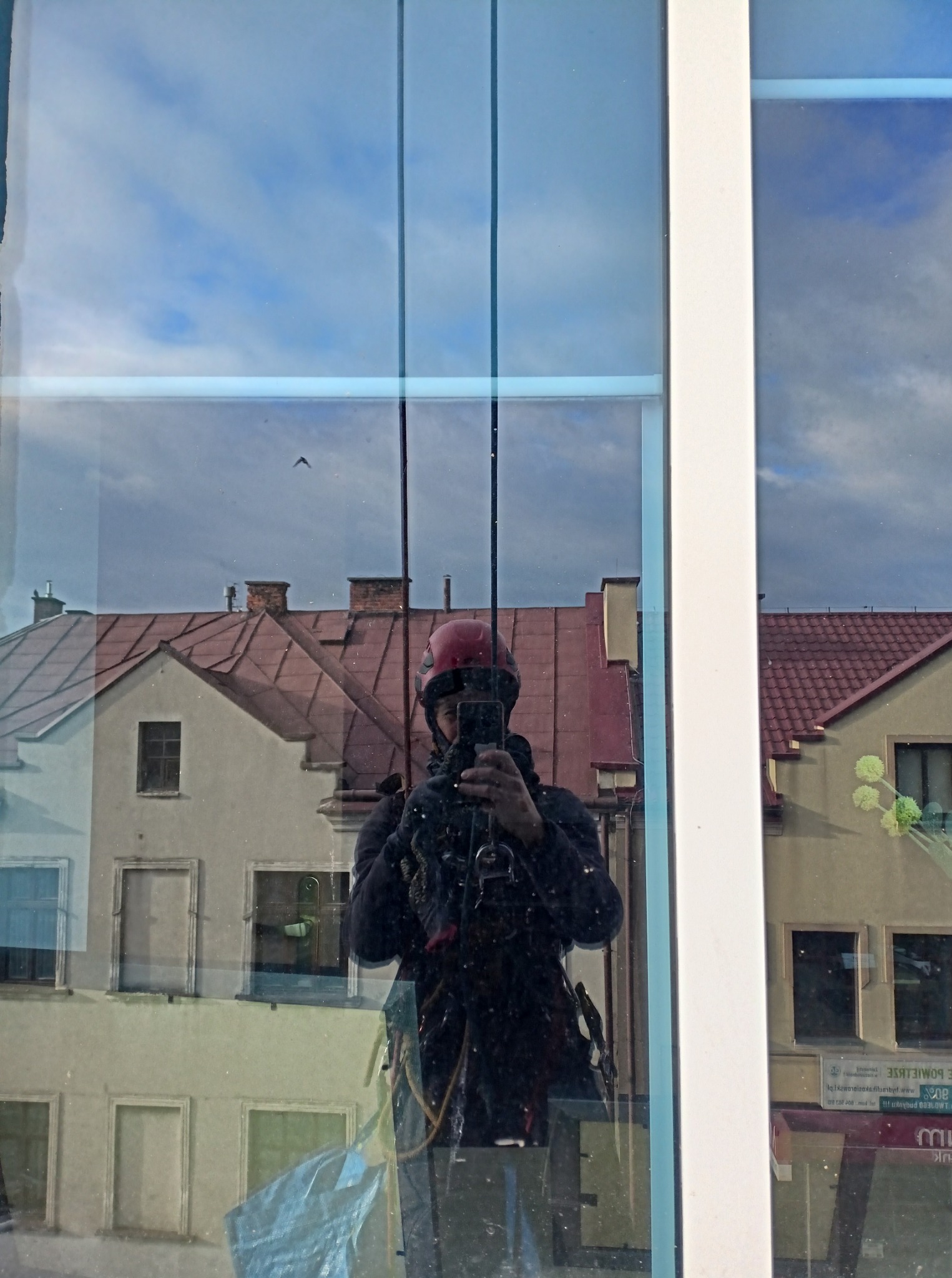 Pracownik w kasku, zabezpieczony linami, robi zdjęcie smartfonem w odbiciu okna budynku. Widoczne budynki w tle i niebo.