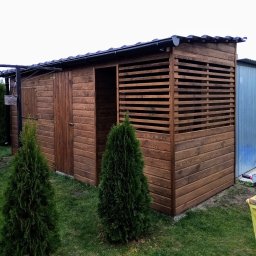 Tomasz Urban - Drewniana konstrukcja z zadaszeniem, częściowo ażurowa, w ogrodzie. Widoczne drzwi i elementy dekoracyjne. Całość w ciepłym, brązowym odcieniu.