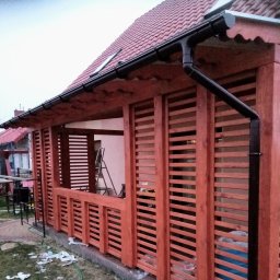 Tomasz Urban - Zadaszona konstrukcja z drewna z lamelami, widok z zewnątrz. Czerwona dachówka na budynku w tle. Elementy konstrukcyjne z wyraźnym rysunkiem drewna.