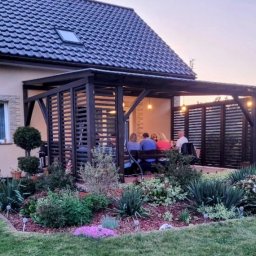 Tomasz Urban - Ciemna pergola z drewnianymi lamelami, oświetlona lampkami, z grupą osób siedzących w środku. Przed pergolą zadbany ogród z kwiatami i krzewami.