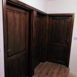 Tomasz Urban - Trzy drewniane drzwi w ciemnym odcieniu brązu, z widoczną strukturą drewna, zamontowane w białej ścianie, z fragmentem podłogi imitującej drewno.