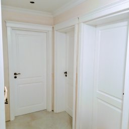 Tomasz Urban - Wnętrze z trzema białymi, panelowymi drzwiami z ciemnymi klamkami. Widoczny fragment sufitu z listwami dekoracyjnymi i mały pies rasy szpic miniaturowy.