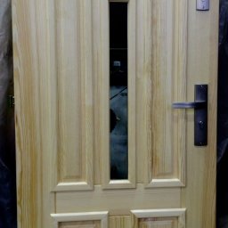 Tomasz Urban - Drewniane drzwi wejściowe z szybkami, surowe, bez wykończenia. Widoczne okucia i zamek. Charakterystyczny wzór drewna.