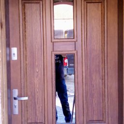 Tomasz Urban - Frontowe, brązowe drzwi wejściowe z szybkami, widoczny srebrny zamek i klamka. W szybach odbija się sylwetka osoby i fragment otoczenia.