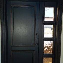 Tomasz Urban - Ciemne, eleganckie drzwi wejściowe z bocznym panelem z trzema okienkami, przez które widać otoczenie na zewnątrz. Klasyczny design z nowoczesnymi akcentami.