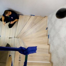 Tomasz Urban - Spiralne schody w trakcie montażu. Widoczny stolarz przy pracy, narzędzia oraz zabezpieczenia na ścianie. Jasne drewno i wzorzysta tapeta w tle.